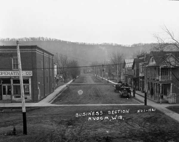 Elevated view of the central business district. The Avoca Cooperative Company is on the street corner on the left. Three men congregate near a car parked at the curb on the right. Another group is gathered behind them on the sidewalk.