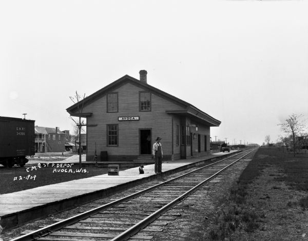 View across railroad tracks of a man and a dog posing on the platform in front of the Chicago, Milwaukee, and St. Paul depot. In the background on the left is a railroad car, and buildings in the central business district.