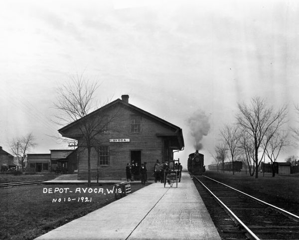 View down platform of people waiting outside the train depot. A train is approaching the terminal on one set of tracks, and a number of train cars are on another track in the background.