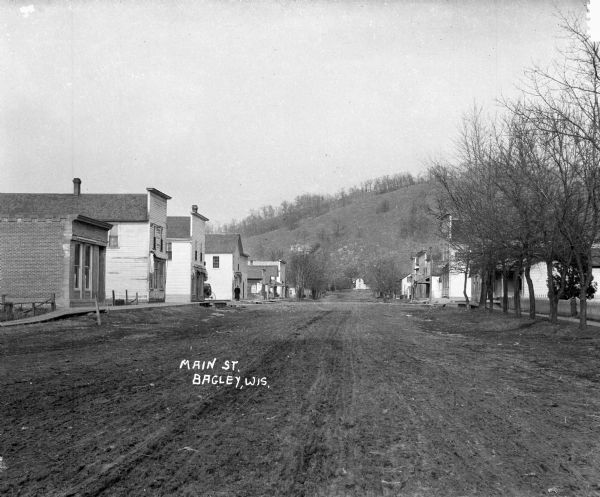 View down unpaved Main Street. A row of houses are on either side of the road, and there is a steep hill in the background.
