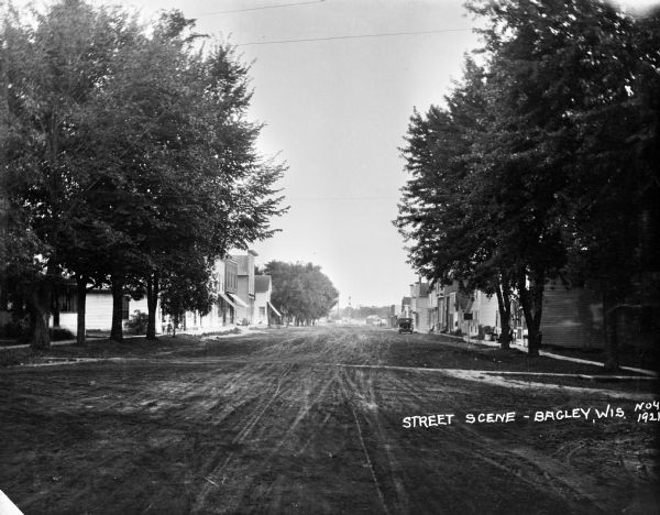 View down a tree-lined dirt road. Houses and shops line both sides of the road, and men and children are sitting and playing on the sidewalks. In the far distance are railroad signals.