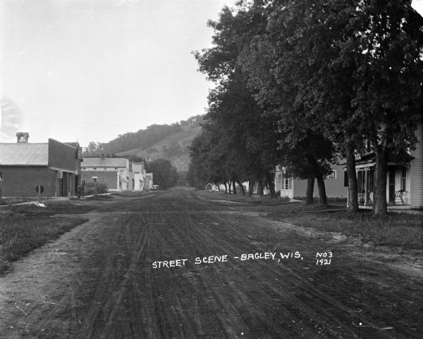 View down dirt road line with trees and houses on the right side. On the left side are commercial buildings, and in the far background is a steep hill.