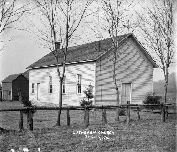 Lutheran church with a fence and trees in front. There is a shed with a ladder propped against it behind the church.