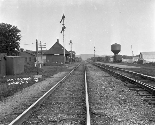 View down railroad tracks looking towards the Bagley train station. On the right is a large storage tank. A man is driving a car near the railroad tracks near railroad cars.