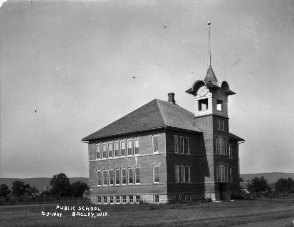 Exterior view of a two-story public school with a tall steeple surrounded by a lawn. In the background are dwellings among trees, and hills in the far background.