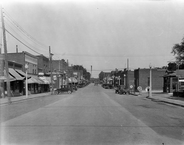 View down street with storefronts on both sides. On the left, there is a tailor shop, Pundy's Cafe, Dubow's[?] Variety Store, a clothing store, and Vander Bie's Ice Cream. On the right, there is a Phillips 66, a jeweler, a meat market, a drug store, and a hardware store, among others.
