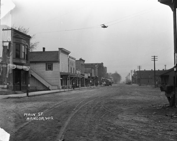 View down Main Street dirt road. A horse and buggy stand near a sidewalk on the left near a group of men. Further down on the right the Bangor State Bank has two horse and buggies stationed in front.