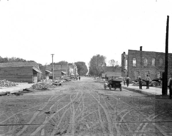 View down dirt road with destroyed buildings on both sides. There are many piles of rubble, and some buildings are gutted shells. Pedestrians walk along road and the sidewalk, and a man is driving a car down the road.