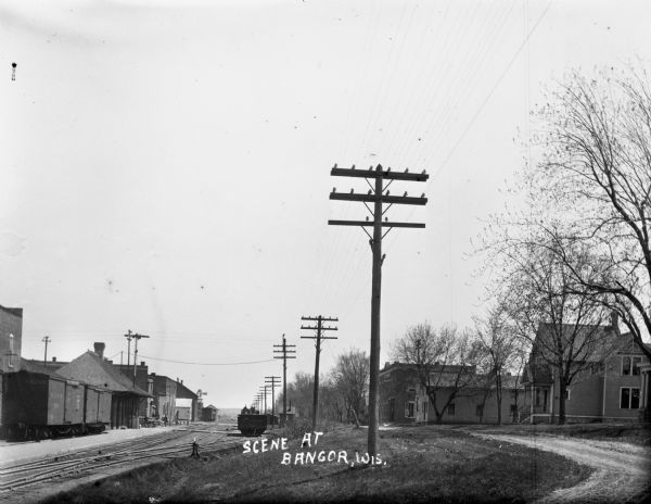 View of railroad tracks near a railroad station on the left and a residential road on the right. There is a row of utility lines leading into the far distance.