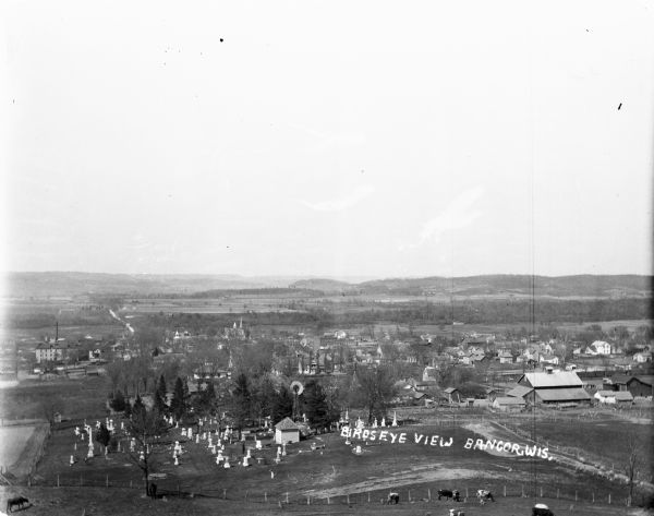 Elevated view of the town with hills in the distance. In the foreground are cattle inside a fence near a cemetery with a windmill. The town is behind further down the hill.