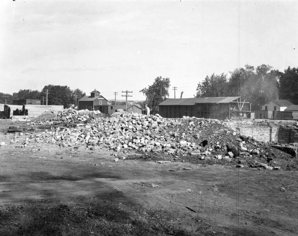 View of a man standing behind a pile of rubble. In the background is a barn, railroad car, and a large open shed stacked with lumber. Part of a foundation can be seen on the right.