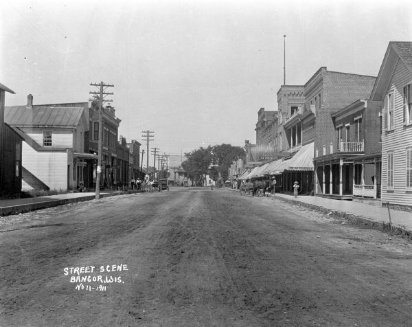 View down dirt road. There is a millinery, dentist's office, drug store, Elsen House ice cream, and meat market. There are horse-drawn carriages on either sides of the street, and pedestrians on the sidewalks. A gazebo is in the distance.