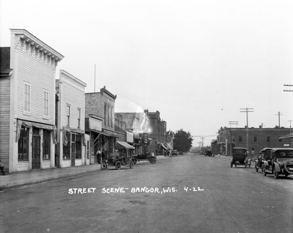 View down a street, where automobiles are parked along both sides of the road. Pedestrians are on the sidewalk on the left in front of storefronts and the Farmers State Bank.