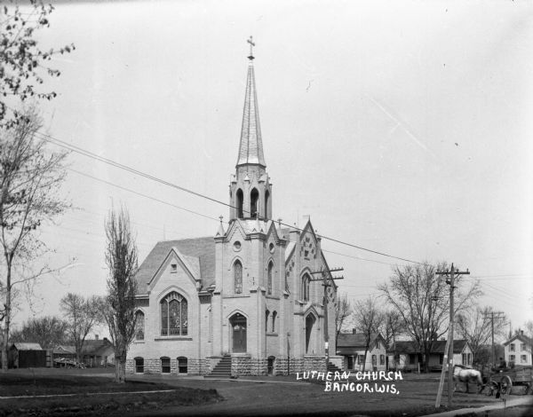 Exterior view across road of Lutheran church with stained glass and a steeple with a bell and cross. A horse and carriage are moving down the street on the right.