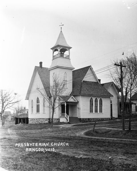 View across road towards a church with a steeple that features a bell. There is an open shed behind the church, and houses are in the background. A datestone on the side of the church reads: "AD 1884". Caption reads: "Presbyterian Church Bangor, Wis."