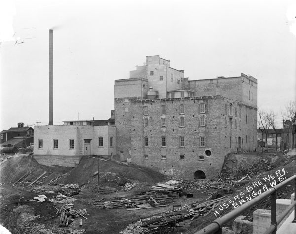 Elevated view of back of Hussa's brewery. There is a tall chimney on the left, and a yard with piles of lumber is in the foreground.