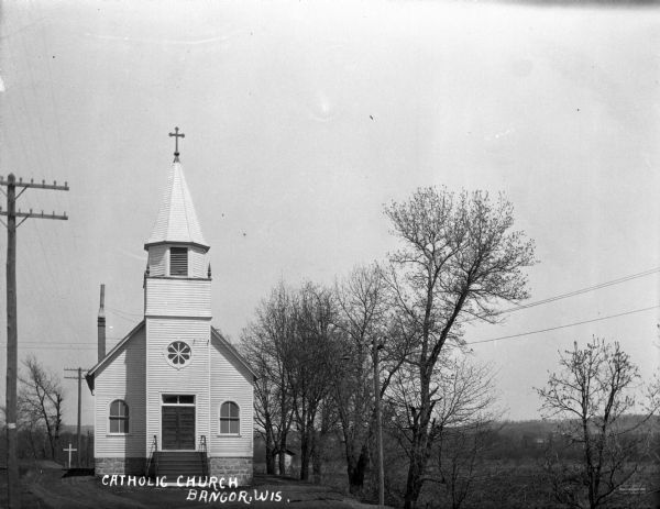 View of front exterior of church with a steeple with a cross on top. On the right is a row of trees, and beyond there appears to be a field or marshy area. In the background are low hills.