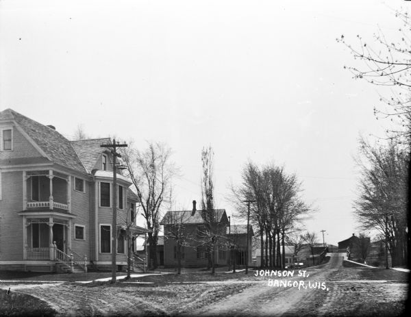 View down Johnson Street. There is a two-story house on the corner on the left. A row of power lines and trees line the street to a bridge in the middle distance. On the far side of the bridge is a barn.