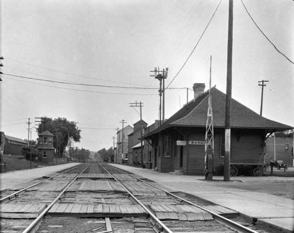View down railroad tracks. On the left, there is a two-story, octagonal hut. On the right, there is a railroad station with a Western Union sign. Industrial buildings are in the background.