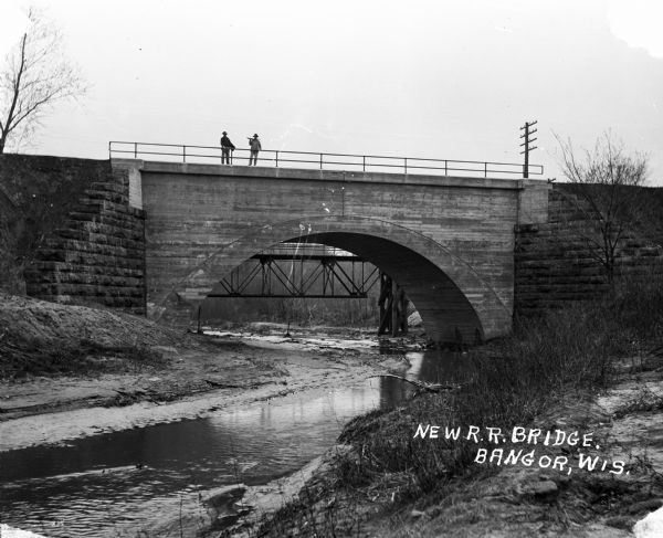 View from shoreline of two men standing on a newly constructed railroad bridge over a stream.
