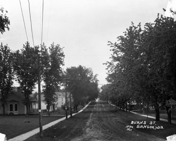 View down tree-lined Burns Street, which is on a hill. Houses and sidewalks are on both sides of the street, and a small group of men are in the yard of a house on the left.