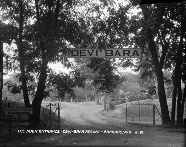 View of the entrance to the Devi-Bara Resort. There is sign on the fence that reads, "Saddle Horses - 1/2 block" and another sign on a tree denoting that this is an American Automobile Association (AAA) official resort. Past the entrance, there is a parking lot and outdoor seating.