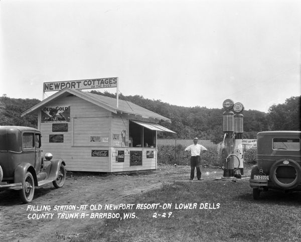 Filling station with attendant standing outside near two pumps. There is a stand with signs advertising Old Gold cigarettes, 5 cent vegetable sandwiches, Prima Special, Hires, and Coca-Cola. A clerk is standing inside the filling station stand. A sign on the roof advertises "Newport Cottages on the River."
