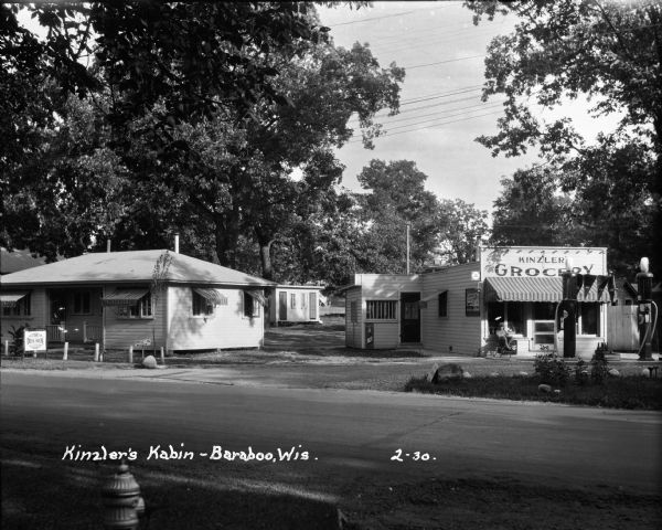 View from across road of a grocery store and gas station, with a home next door. The store features two pumps and advertises Deep Rock motor oil, Coca-Cola, and Pabst Blue Ribbon Beer. There is a man leaning back in a chair under an awning at the front of the Kinzler grocery store.