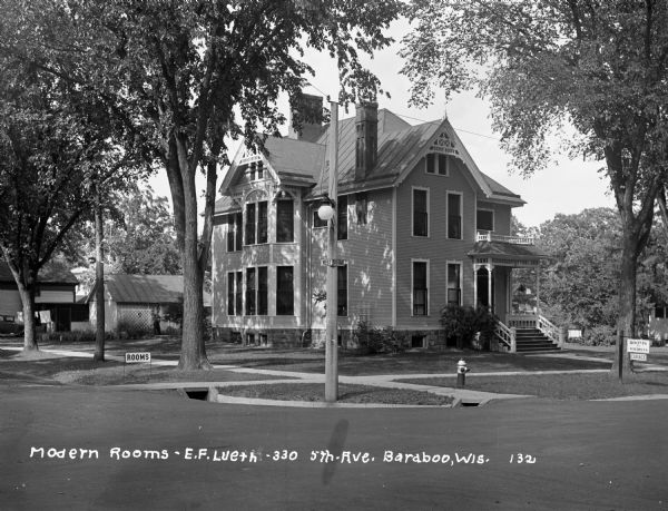 View across road of a two-story home located on a corner lot at 330 5th Avenue. There are signs on the lawn advertising rooms for tourists. A man is standing in the backyard.