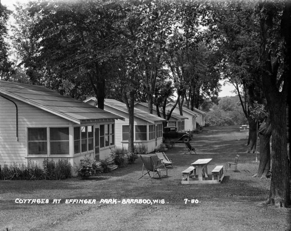 Row of cabins in Effinger Park. Each has a screened back porch. Picnic tables, sling-back chairs, and a parked car are under the trees on the lawn.