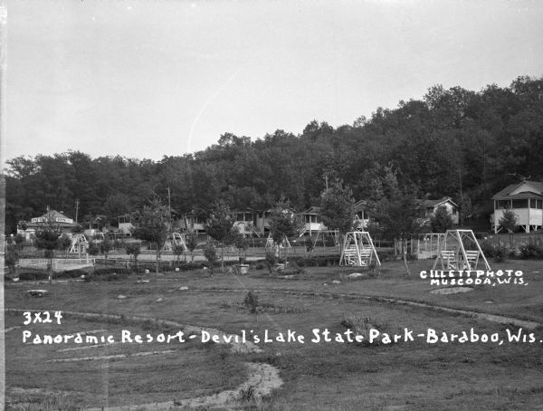 View of two-seat lawn swings amid trees on a lawn. Cabins are along a hill in the background. There is an information center on the far left.
