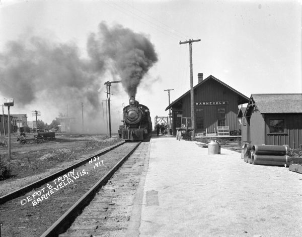 View of a train approaching the depot. Two men stand alongside the train. Heavy smoke from the train causes a fog cloud.