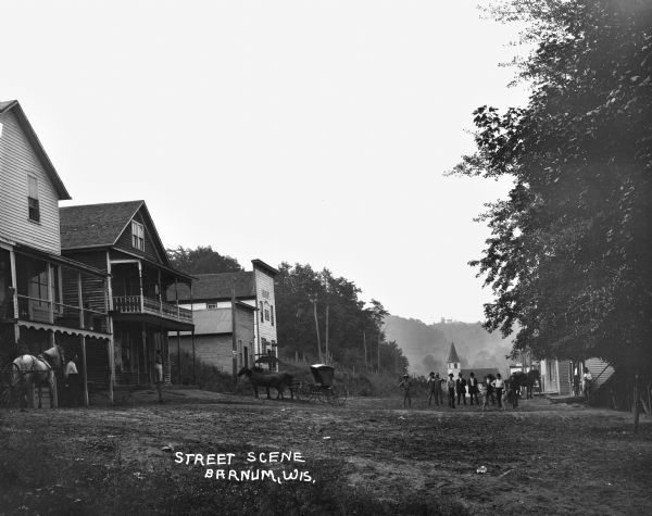 Group of men and children standing in the middle of the unpaved street. On the left are storefronts, and in the far distance is a church and tree-lined ridge.
