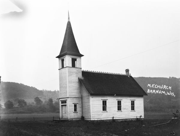 Exterior view of the Church. The building features double doors, a belfry, and a steeple with a cross. There is a cart parked in the yard in the lower-right corner. Hills are in the background.