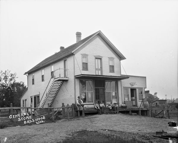 Four men, a dog and a boy sitting on the porch of the general store. Signs in the windows read, "Ferry & Co. Seeds" and "Rape seed for sale." A barn and windmill are in the distance.