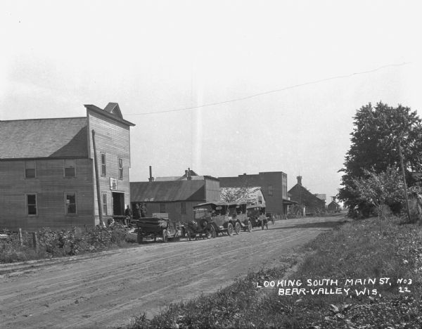 View from roadside of Main Street. There are three men wearing coveralls standing near the open door of a building. Four cars are parked along the street