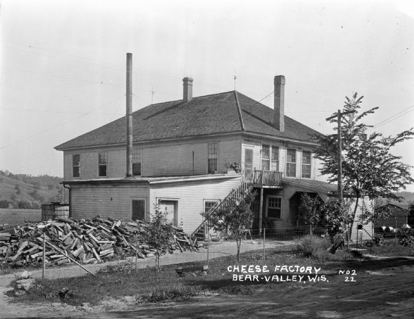 Exterior of a two-story cheese factory. The building features a set of stairs leading to the second-floor entrance and three chimneys. There is a large pile of chopped fuelwood in the yard.