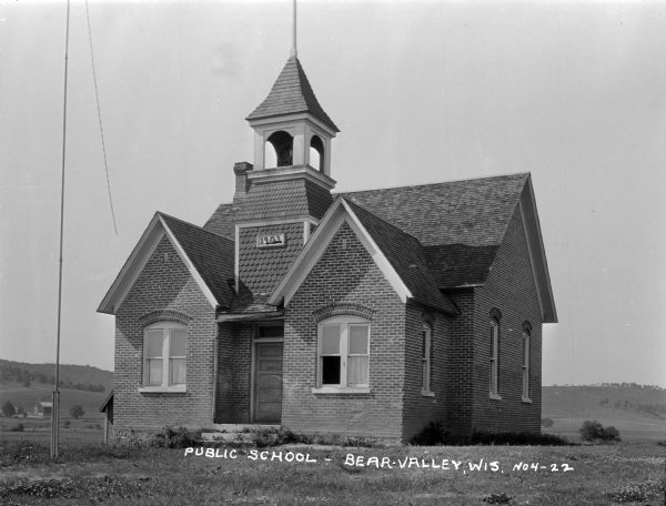 Exterior of a schoolhouse with a bell tower and flag pole. In the background is a farm, and fields and hills. A sign on the side of the tower says "1903".
