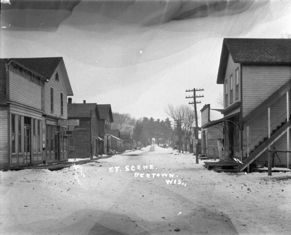 View down a snow-covered street. There are shops on either side. "W.J. Birch - Cash Store" is on the left.
