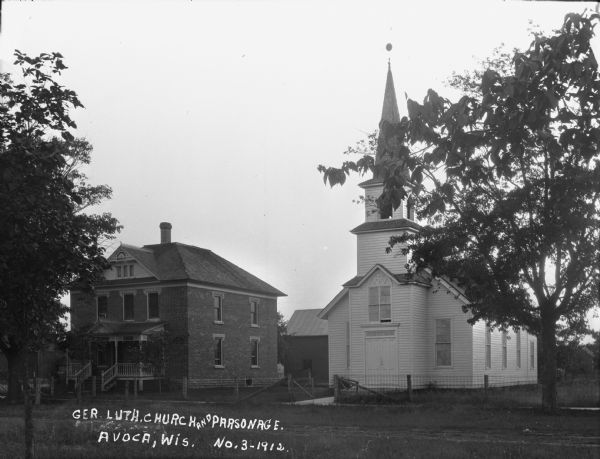 View across street of a Lutheran church and a parsonage next door. The church has a tall steeple with a bell.