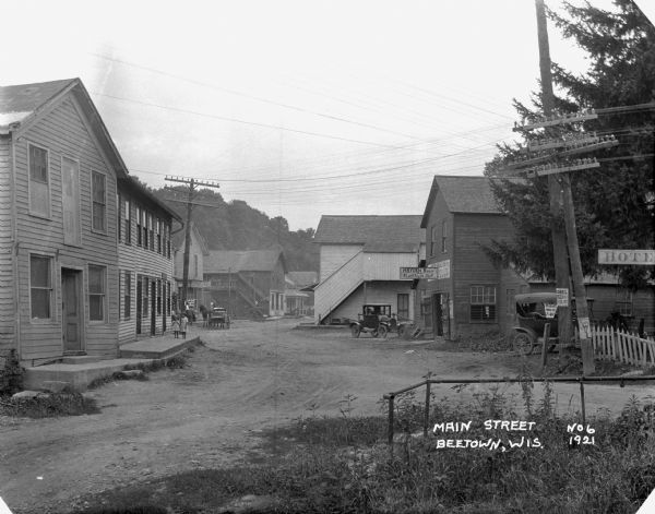 Main Street, where two young girls walk on the sidewalk, and a horse and carriage and two cars are parked in the street. There is a townhouse complex on the left. "Hayden Brothers Blacksmith Shop" and the banner of a hotel are on the right. Electric power lines are along the street.