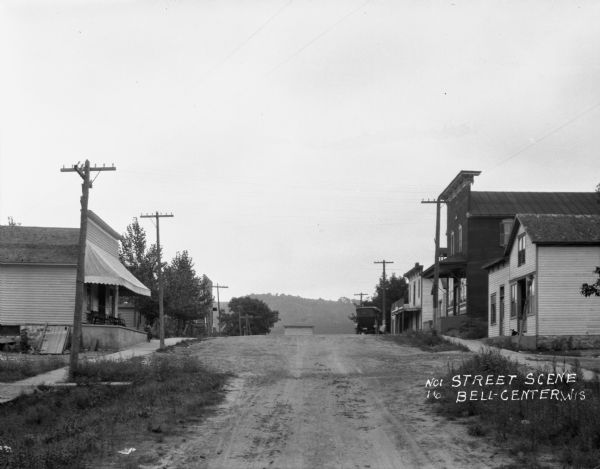 View up the hilly street with shops on either side. A car is parked at the entrance of the hotel on the right. A young boy sits on the porch under an awning of the shop on the left.
