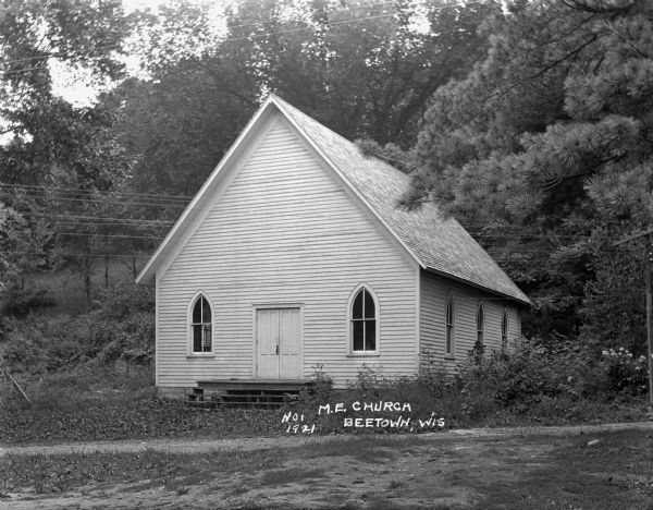 Exterior of the Methodist Evangelical church surrounded by trees. The building features a steps and a small landing at the entrance, arched windows, wood siding, and a steep gable roof.