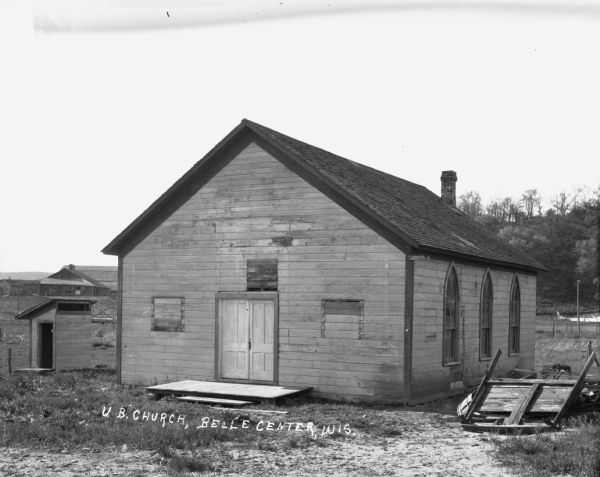 Exterior of the one-room United Brethren [?] Church. There is an outhouse to the left of the church. On the right is a hill with trees. Farm building and a barn are in the distance.