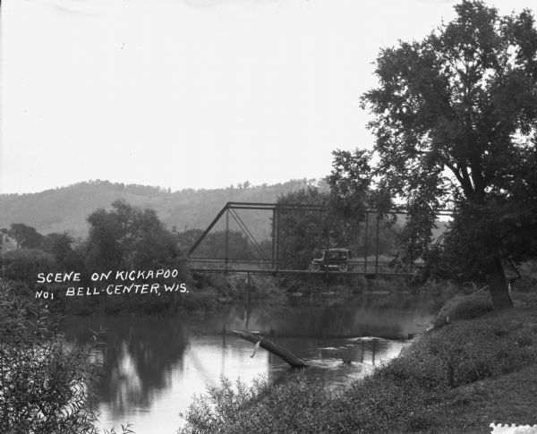 View from shoreline of a car driving over a bridge on the Kickapoo River. There is a ridge of a hill in the background.
