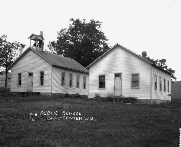 Exterior view of two public school buildings. The left building has a bell tower, with a flag hanging horizontally from peak of the roof over the entrance.