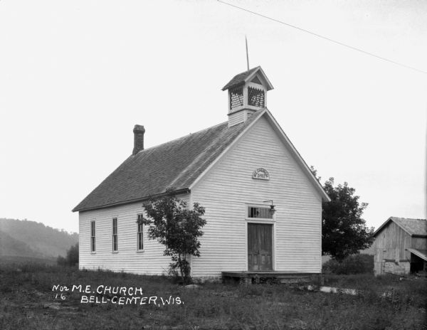 Exterior view of the Methodist Evangelical Church. The building features a sign that says: M.E. 1893. There is a lamp over the entrance, a small, lattice-enclosed bell cupola, and double front doors.