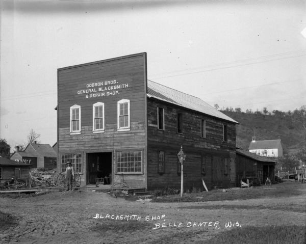 Exterior view from across road of a man in overalls standing in front of the Dobson Brothers General Blacksmith and Repair Shop. The Methodist Evangelical Church is in the middle distance, and a steep hill is in the far background.