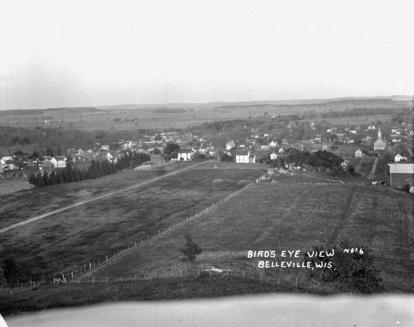 Bird's-eye view of town. Fields, with farms and fences, surround the town made up of numerous dwellings and a church.