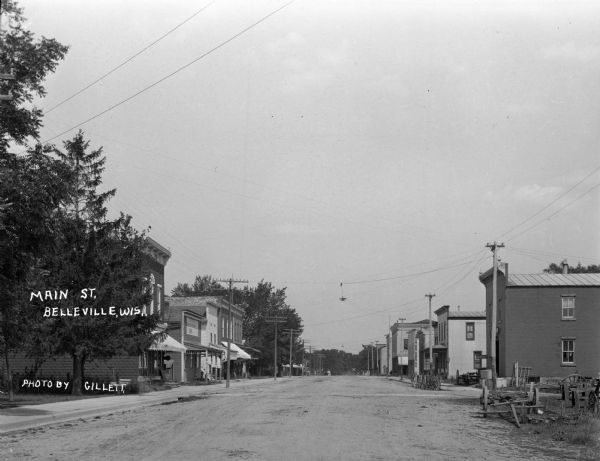 View down Main Street. Rows of shops are on either side. Carriages and carts are parked along the curb. Streetlights hang from the utility lines. A railroad crossing is in the far distance.